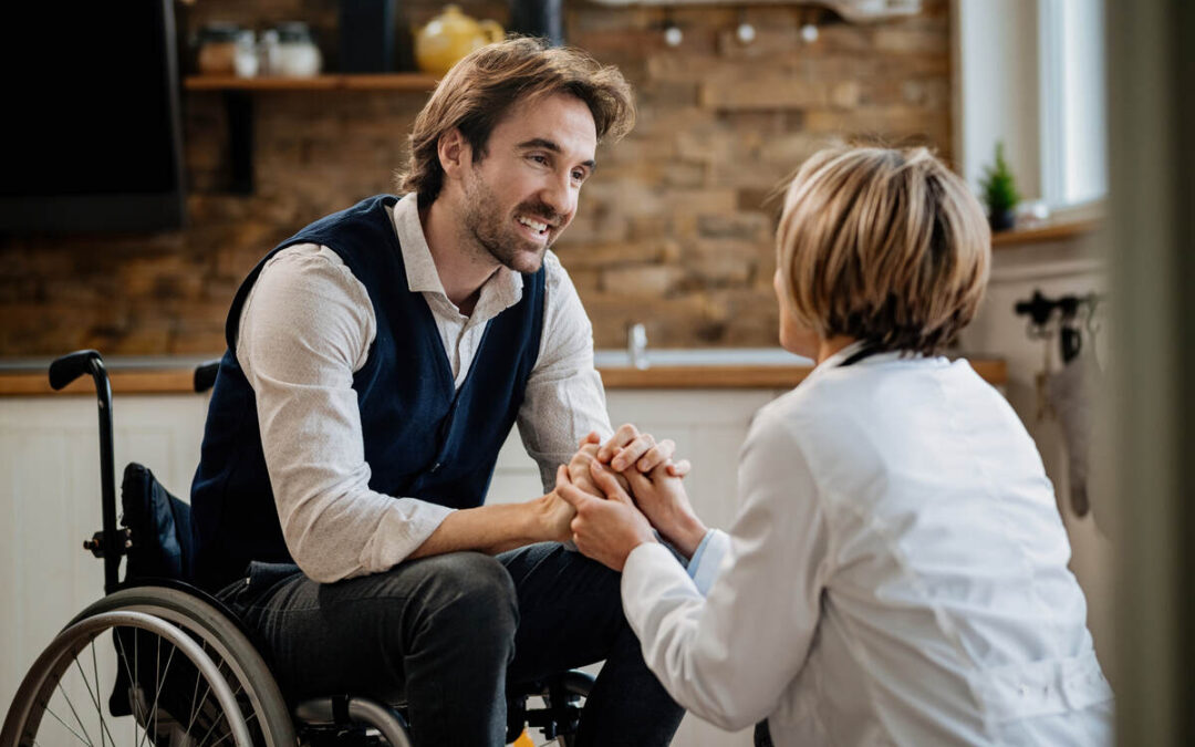 Capability Care NDIS for Community Participation - Young happy man in wheelchair holding hands with his doctor while talking to her during home visit.